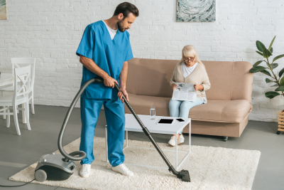 young caregiver cleaning carpet with vacuum cleaner while senior woman reading newspaper on couch